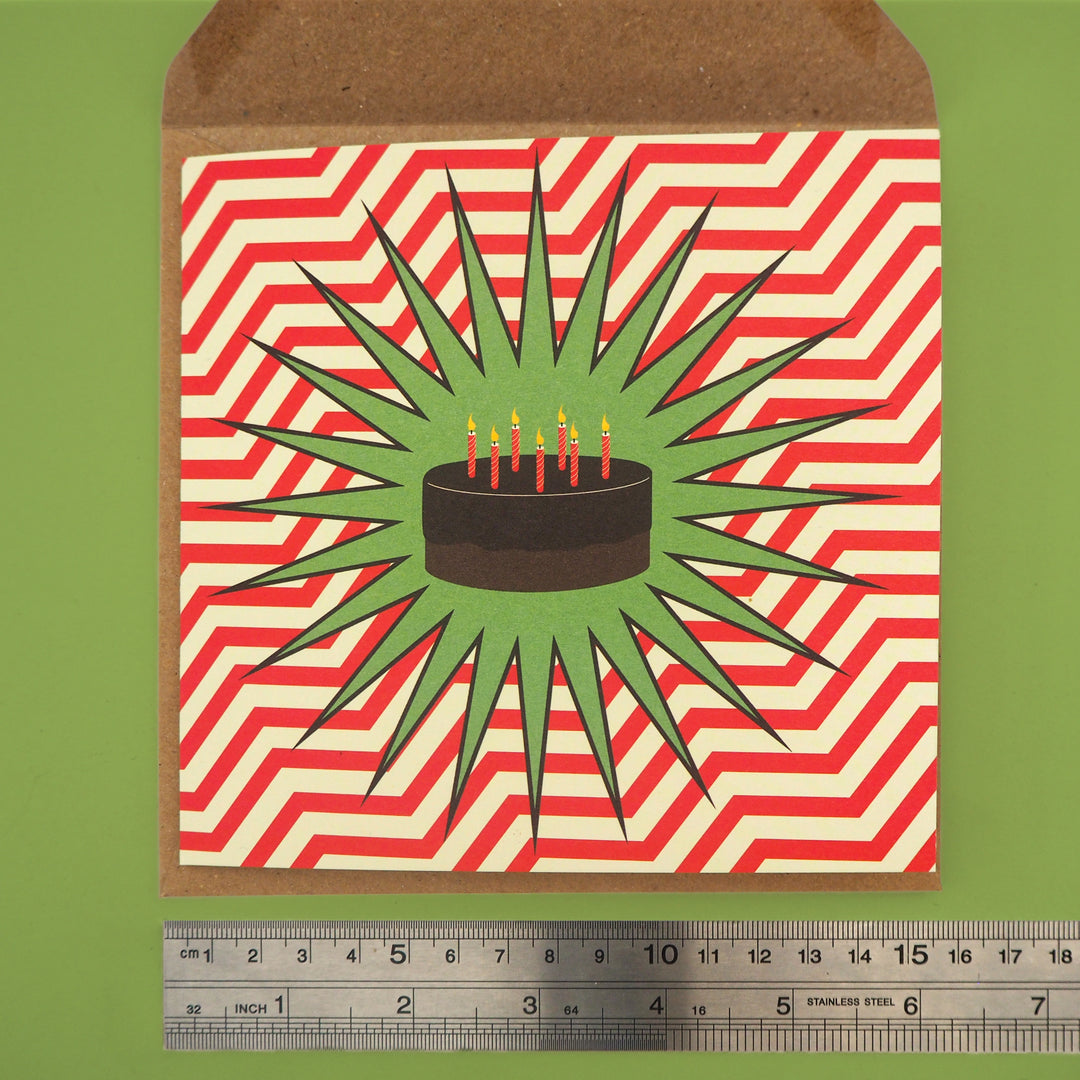 Birthday card on envelope. Card has an illustration of a chocolate cake  and candles on a green star with red and white striped background,. Below there is a ruler for scale.