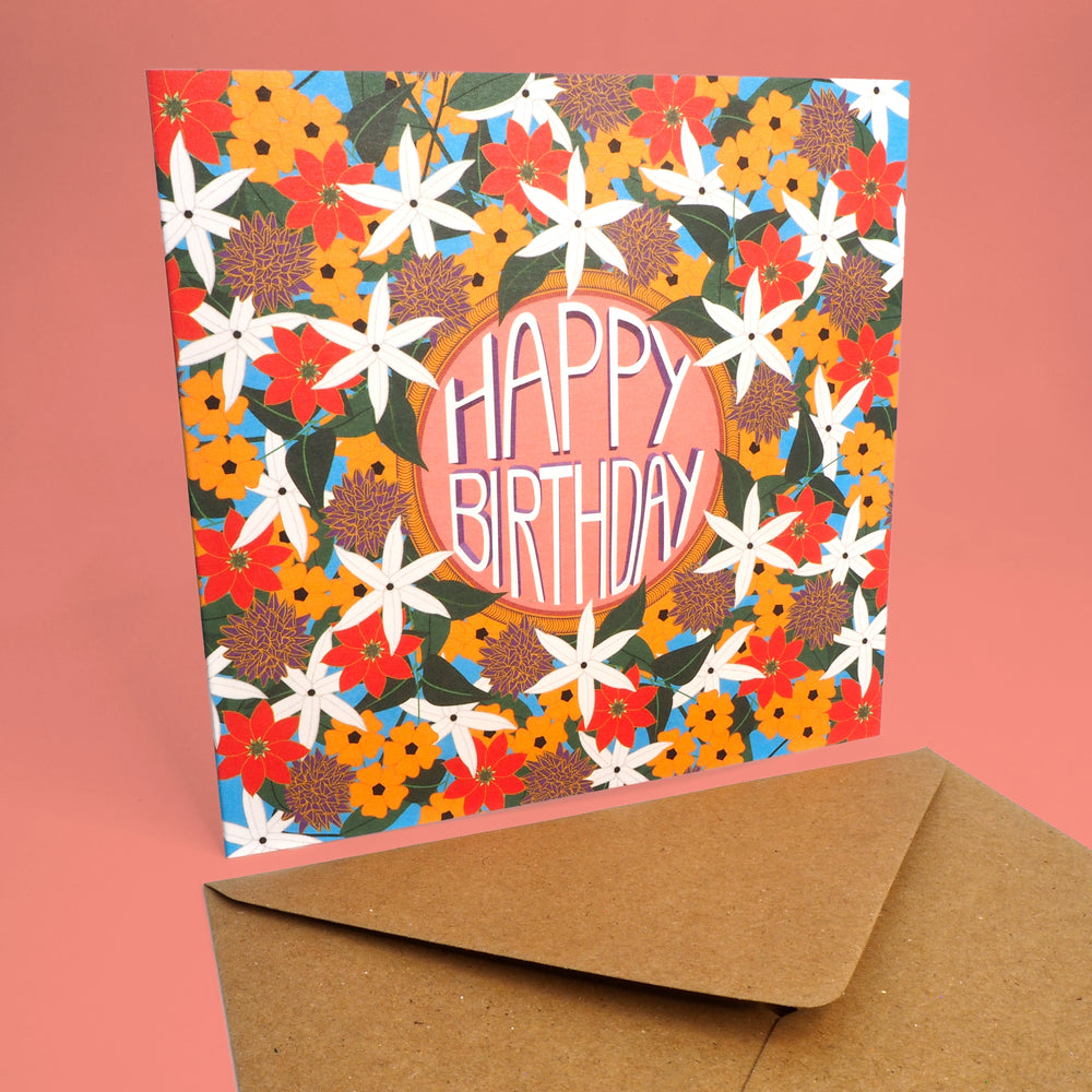 Colorful floral birthday card with 'Happy Birthday' text on a pink background. Stood up with brown craft envelope flat on table
