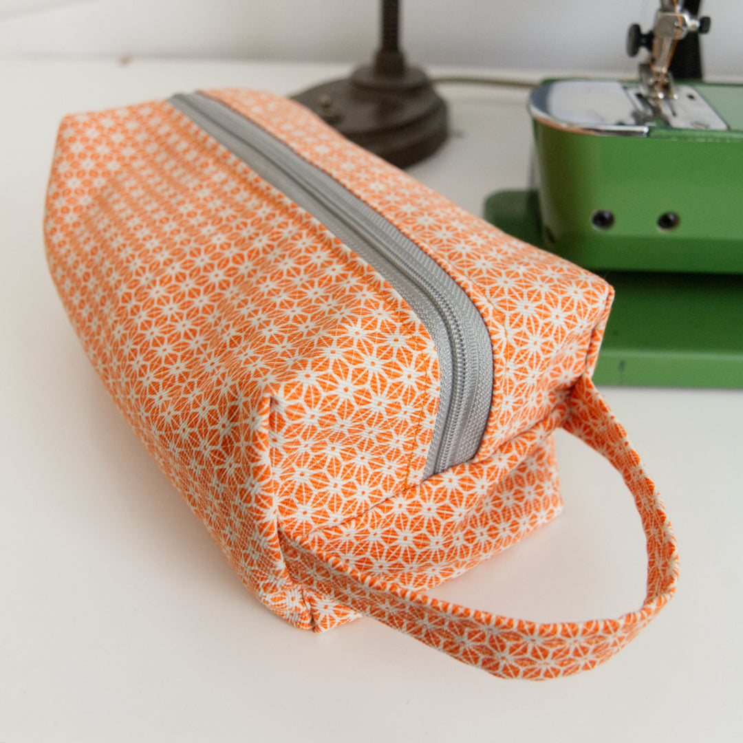 Bright Stem Orange patterned toiletry wash bag with a grey zipper on a white surface with sewing machine in the background