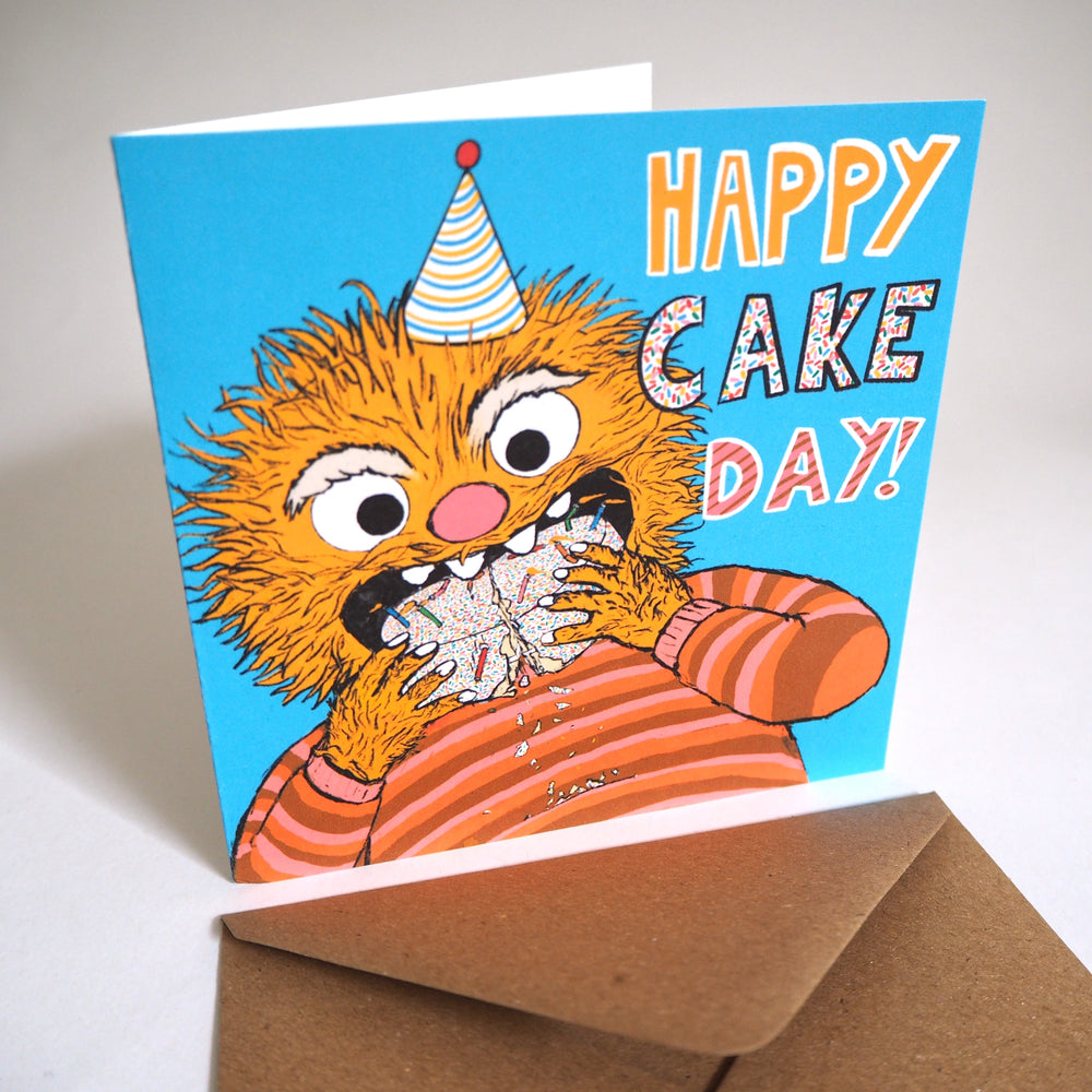 Birthday card with Illustrated character eating a whole cake with candles and 100s  and 1000s falling off. Text saying 'Happy Cake Day!', photographed with a brown recycled envelope.