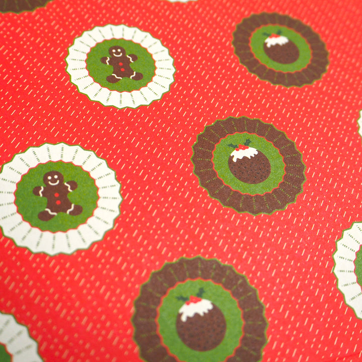 Close up of Bright stem Christmas wrapping paper featuring ginger bread man and Christmas pudding illustrations on a red background.