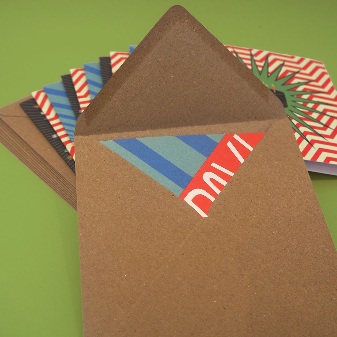 Brown envelope with colorful bright stem birthday card inside. With a stack of birthday cards and envelopes behind all on a green background