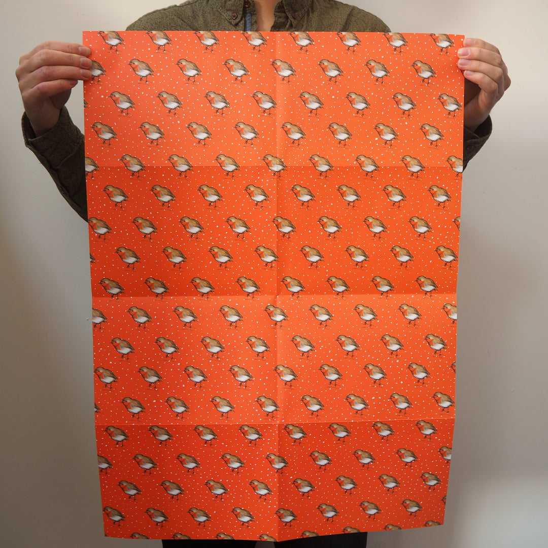 Artist holding a large sheet of bright stem wrapping paper with illustrations of robin bird illustrations on a orange background.