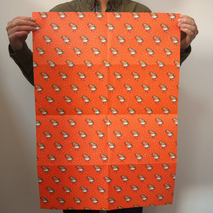 Artist holding a large sheet of bright stem wrapping paper with illustrations of robin bird illustrations on a orange background.