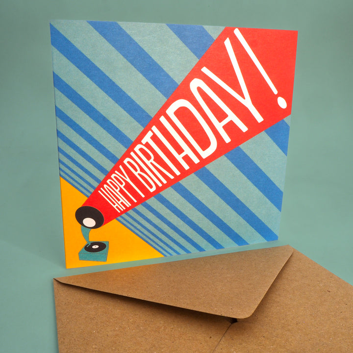 Colourful birthday card with 'Happy Birthday!' text and a record player graphic, accompanied by a brown envelope on a green background. Inspired by the Bauhaus graphic design 