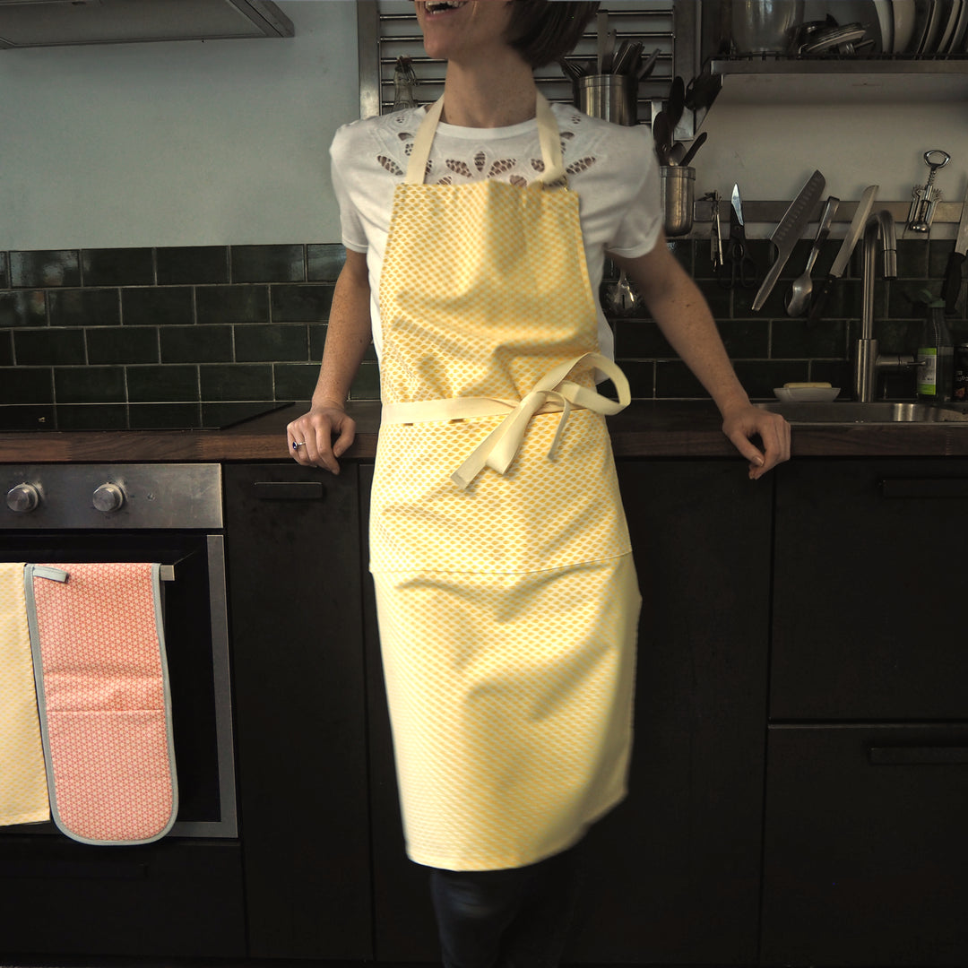 Model wearing bright stem adult apron in kitchen with oven gloves and tea towel hung on oven door.