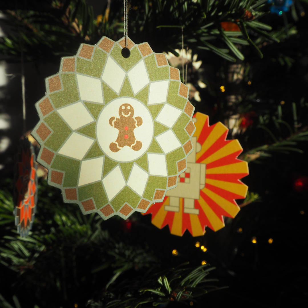 Bright Stem Cardboard Christmas Baubles. Gingerbread man and Robot Designs hung on Christmas tree. Vintage style designs. recycled and recyclable. sustainable design, made in the UK. Held by artist.